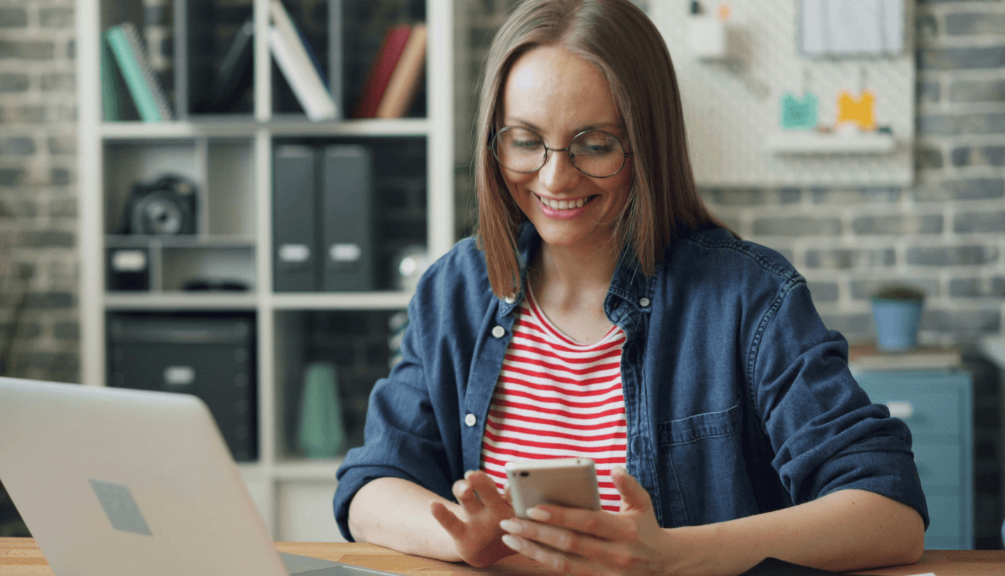 Bild zeigt Frau mit Smartphone in der Hand am Schreibtisch sitzend, vor ihr ist ein offener Laptop.